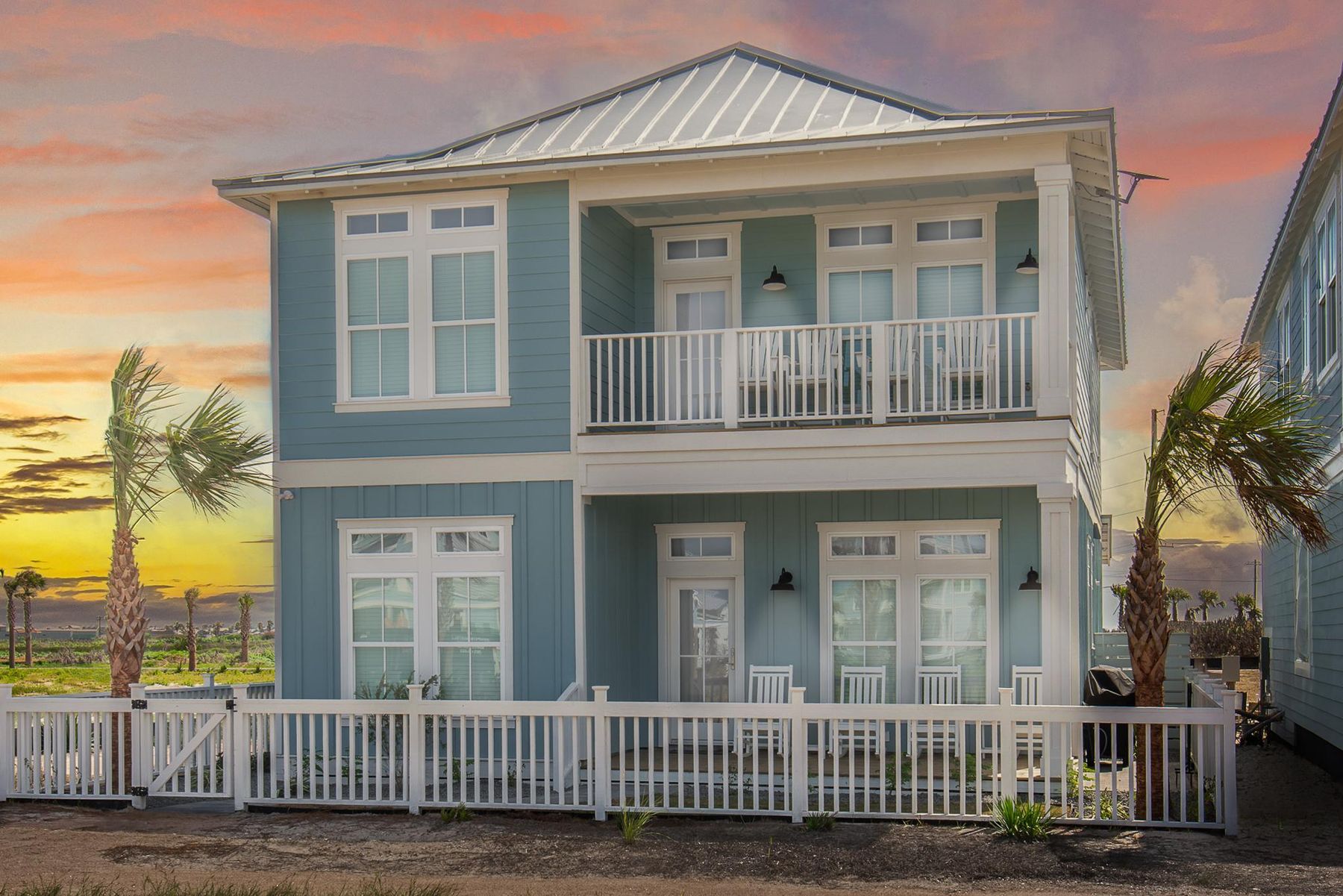 A sea-glass blue coastal home with white porches and a picket fence at sunset
